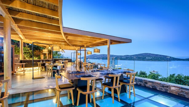 An outdoor dining area with tables and chairs, overlooking a body of water and hills. - Royal Marmin Bay Boutique & Art Hotel - Adults Only (Agios Nikolaos)