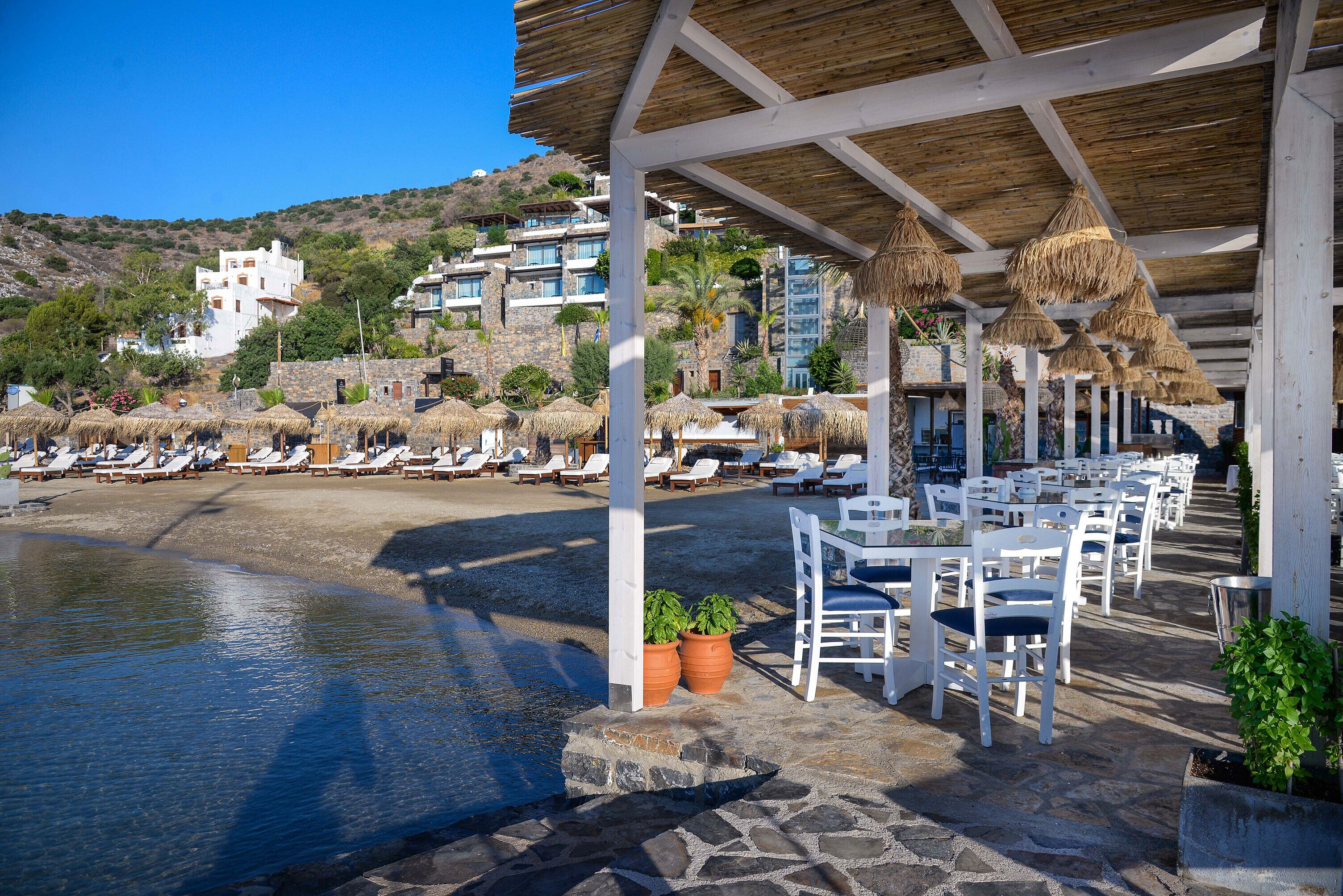A seaside restaurant with outdoor seating, stone flooring, and thatched umbrellas.
