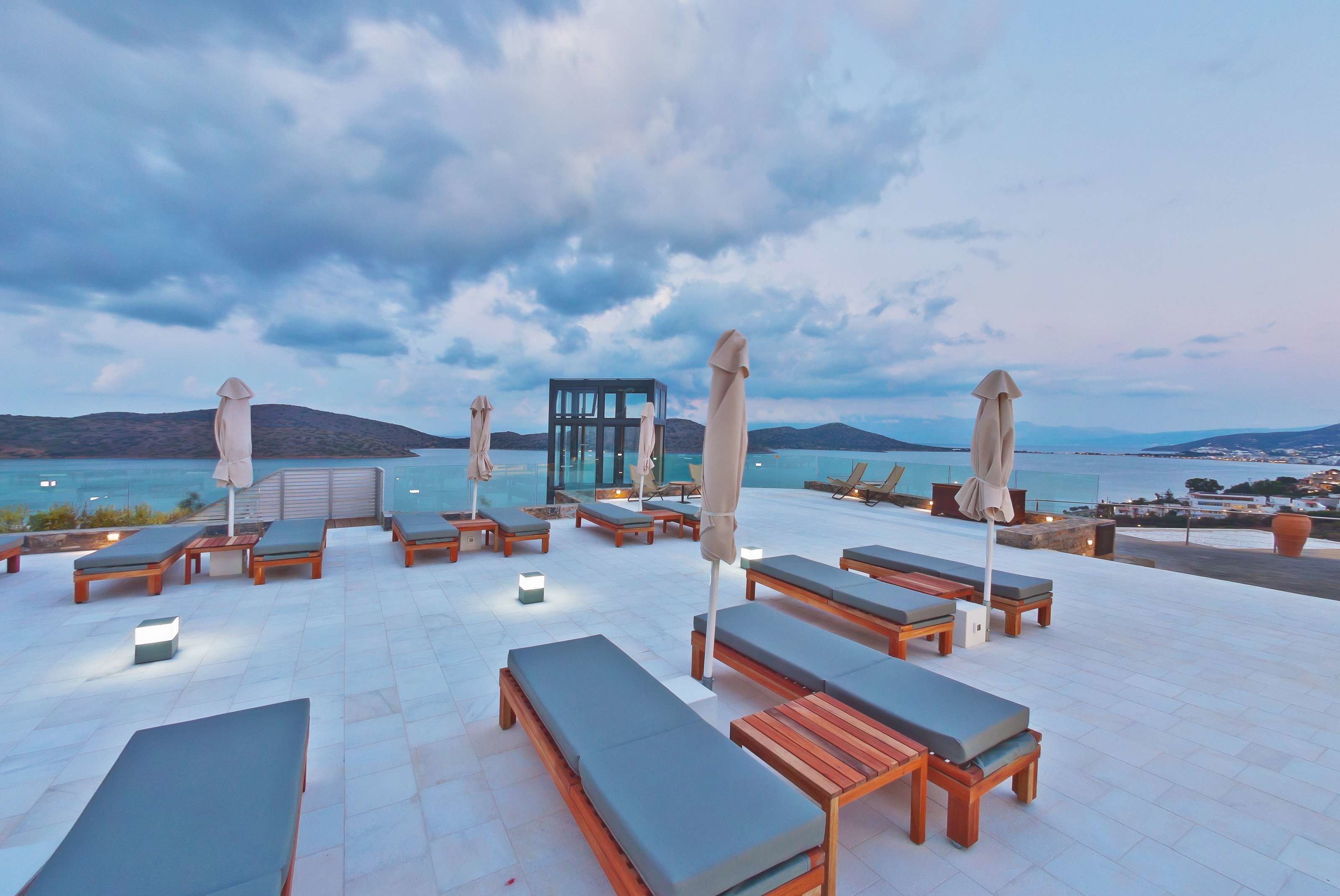 A rooftop terrace with lounge chairs and umbrellas, overlooking a body of water and distant hills.