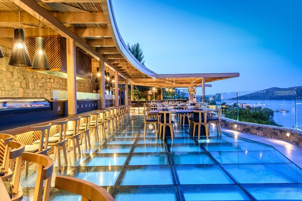 A restaurant with a glass floor, wooden chairs, and tables, overlooking a body of water. - Royal Marmin Bay Boutique & Art Hotel - Adults Only (Agios Nikolaos)