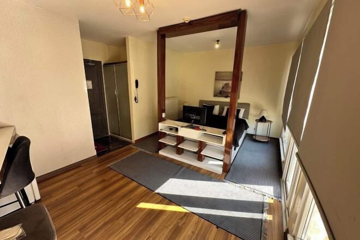 A compact room with a wooden floor, a desk, a chair, a bed, a TV, and a small table with a lamp.