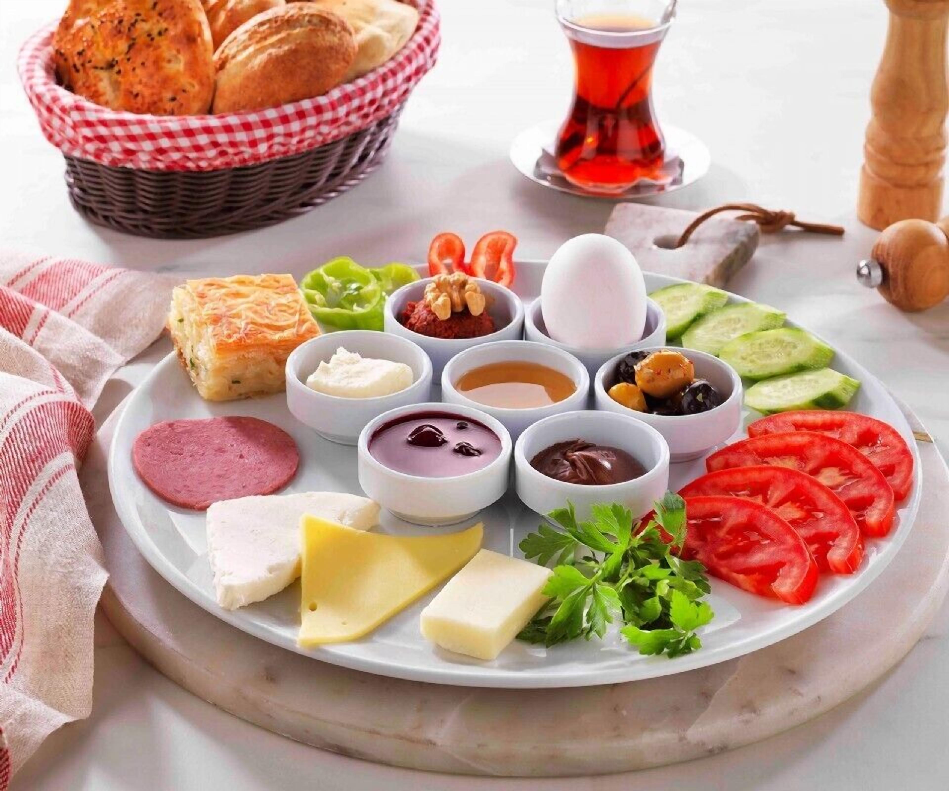 A plate of assorted cheeses, meats, and vegetables, accompanied by a glass of tea and a basket of bread.