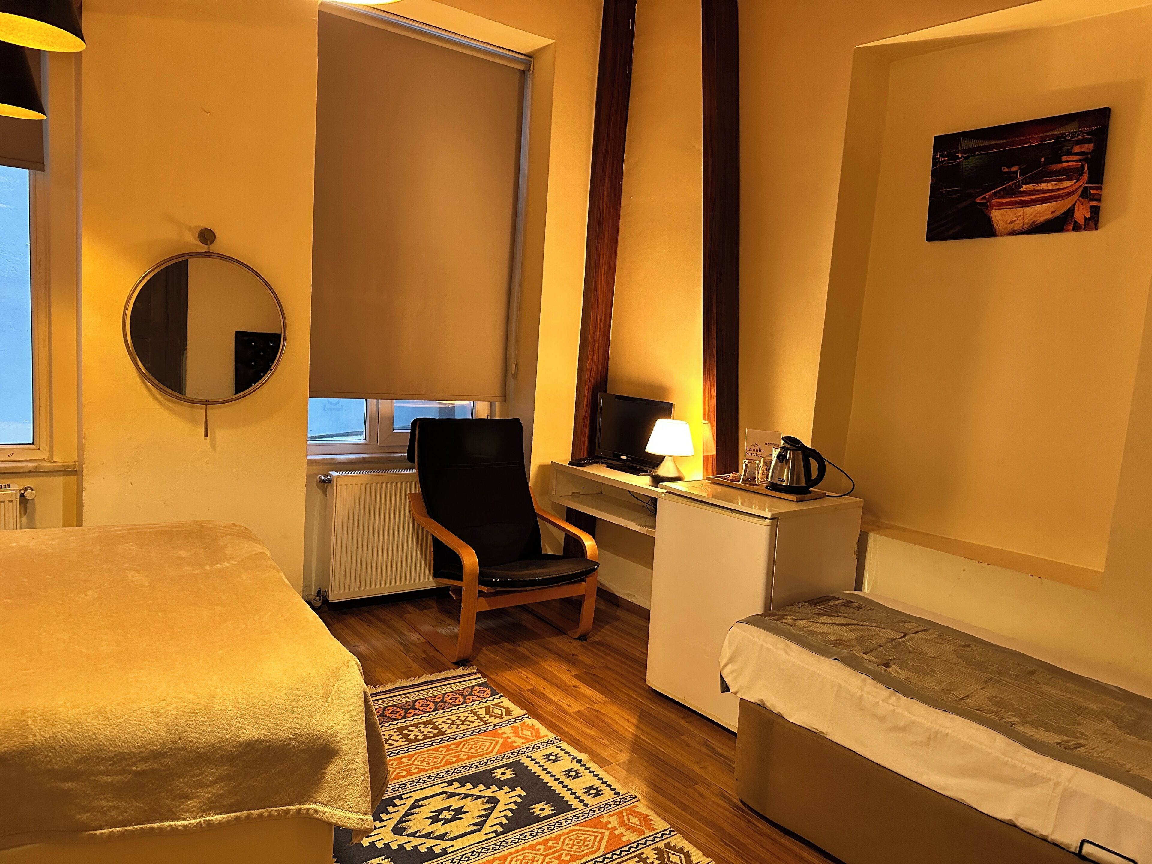 A compact hotel room with a bed, a desk, a chair, a radiator, a window with blinds, a mirror, and a painting on the wall.