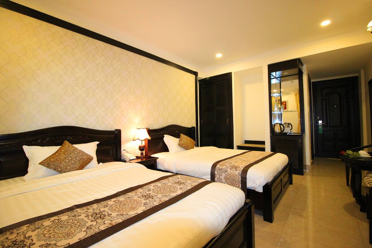 A hotel room with two beds, a nightstand with a lamp, a mirror, and a small table with a vase.