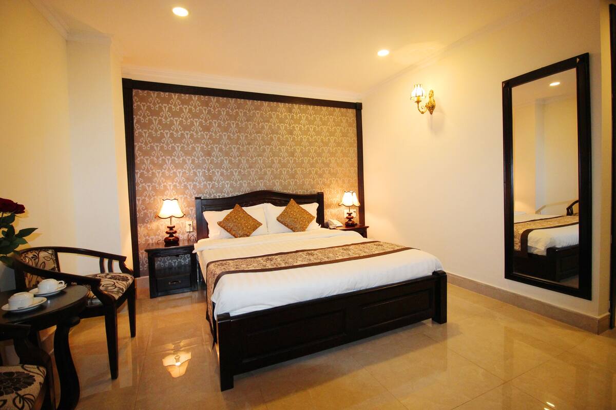 A hotel room with a large bed, bedside lamps, a small table with a chair, and a mirror on the wall.