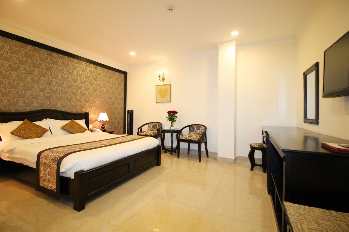 A hotel room with a large bed, a desk, a chair, a small table with a vase of flowers, and a television on the wall.