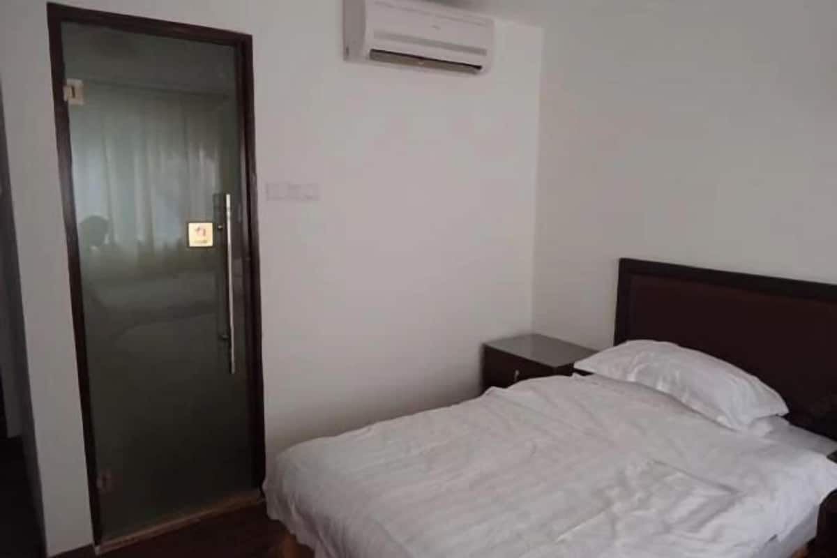 Una habitación con una cama, una mesita de noche pequeña, una puerta con panel de vidrio y un equipo de aire acondicionado en la pared.