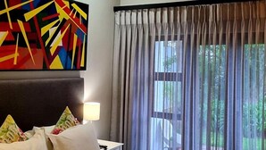 A hotel room with a bed, pillows, a colorful abstract painting, a lamp, and a window with curtains. - Richtershuyz Guest House (Pretoria)
