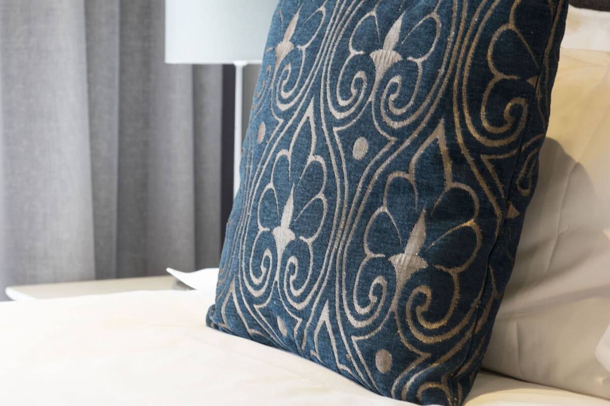 A bed with a blue patterned pillow and a white pillow, a lamp, and grey curtains.
