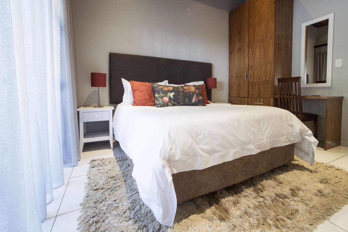 A bedroom with a large bed, a nightstand, a wooden wardrobe, a chair, and a mirror.