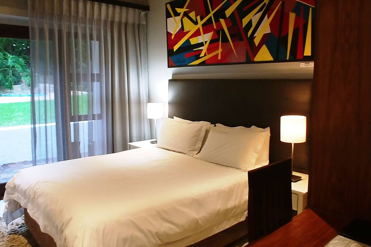 A hotel room with a large bed, two bedside lamps, a colorful abstract painting above the bed, and a view of a garden through the window.