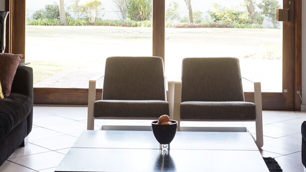 A modern living room with a wooden ceiling, two grey armchairs, a black coffee table, and a view of a garden through large glass doors. - Richtershuyz Guest House (Pretoria)