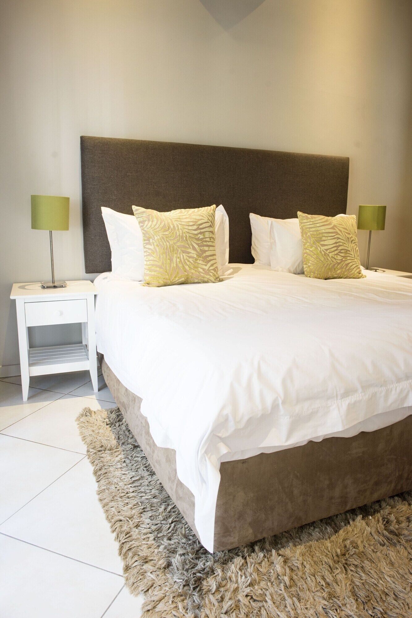 A neatly made bed with white linens and patterned pillows, a brown headboard, and a fluffy rug on a tiled floor.
