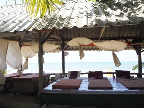 Ein Spa direkt am Strand mit mehreren Massagetischen unter einem überdachten Bereich mit Sonnenschirmen, mit Blick auf das Meer.