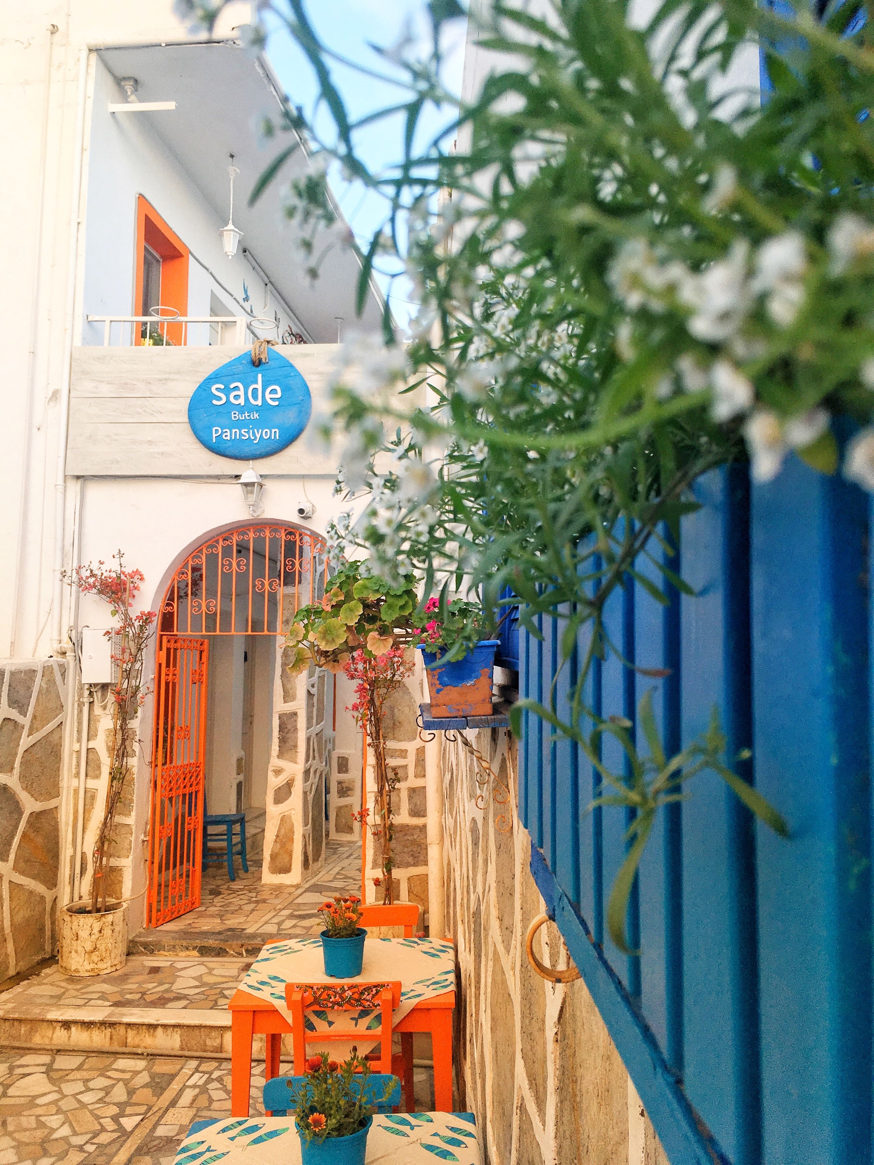 A narrow alleyway with a blue and white building, a sign reading "sade Butik Pansiyon", and a small table with chairs and potted plants.