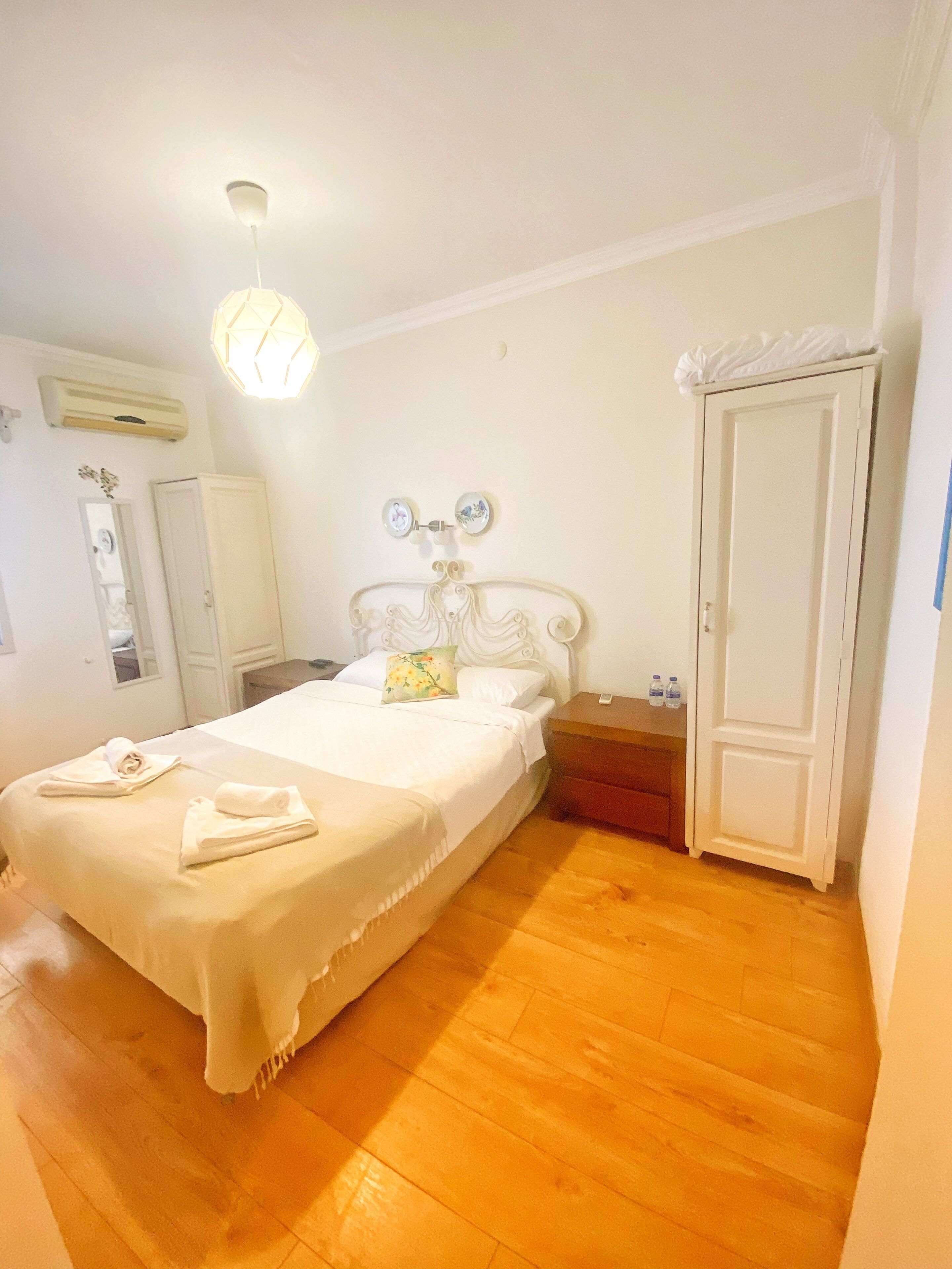Una cama bien hecha con edredón blanco y almohadas, una mesita de noche con lámpara, y un armario en una habitación con suelo de madera y paredes blancas.