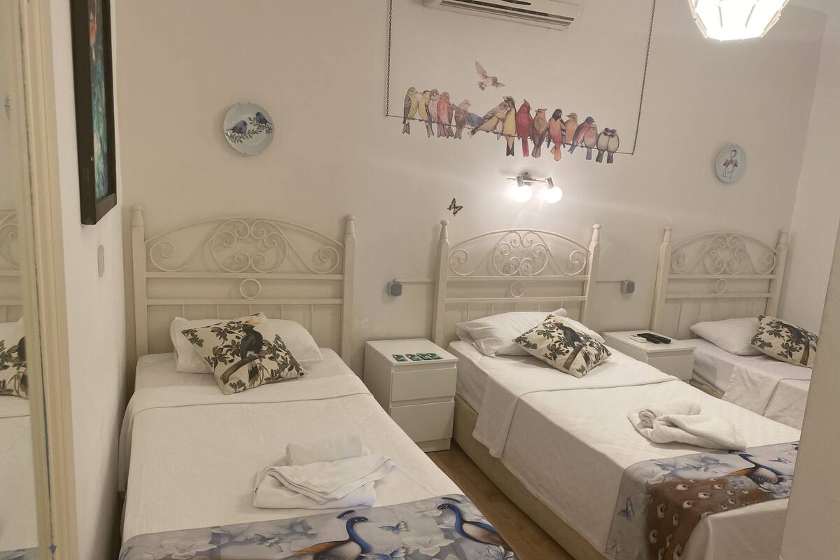Quarto de hotel com três camas de solteiro, cada uma com cabeceira branca e roupa de cama combinando. Há almofadas decorativas, uma mesa de cabeceira com abajur e uma pintura de pássaros na parede.