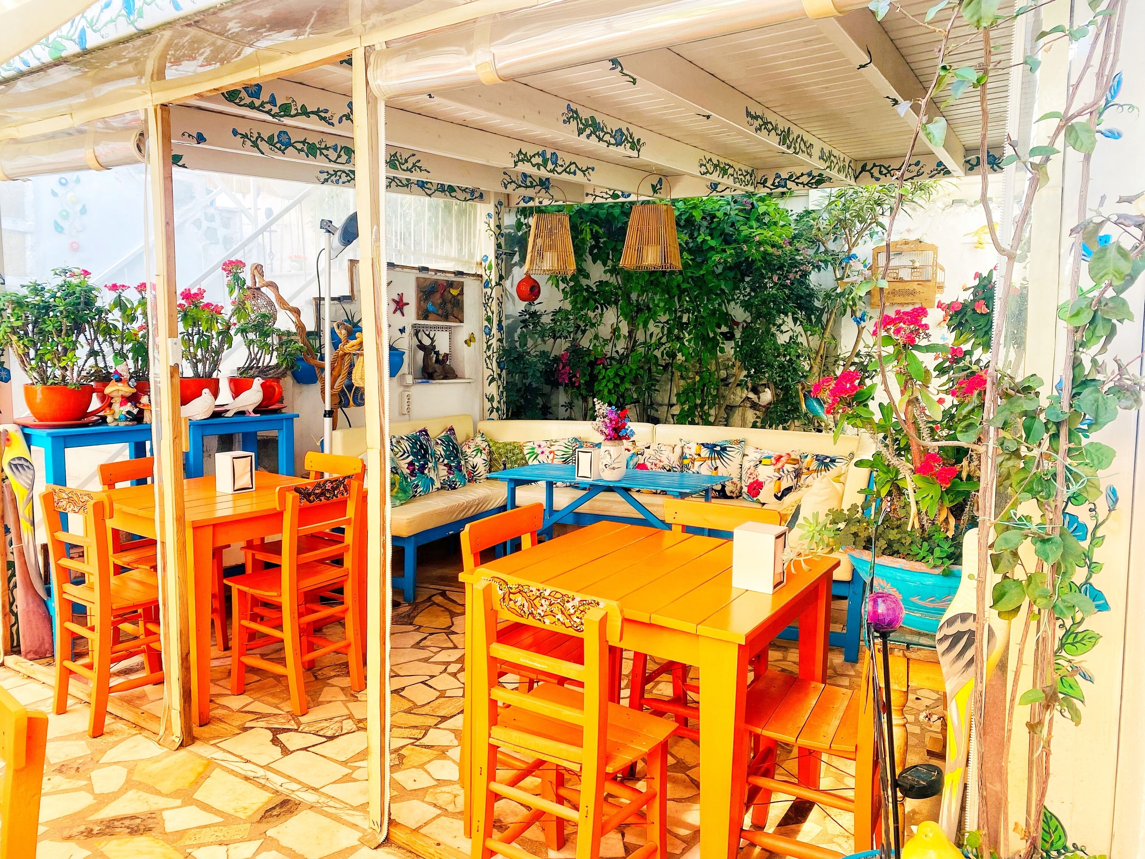 A covered outdoor seating area with wooden tables and chairs, surrounded by potted plants and decorative elements.