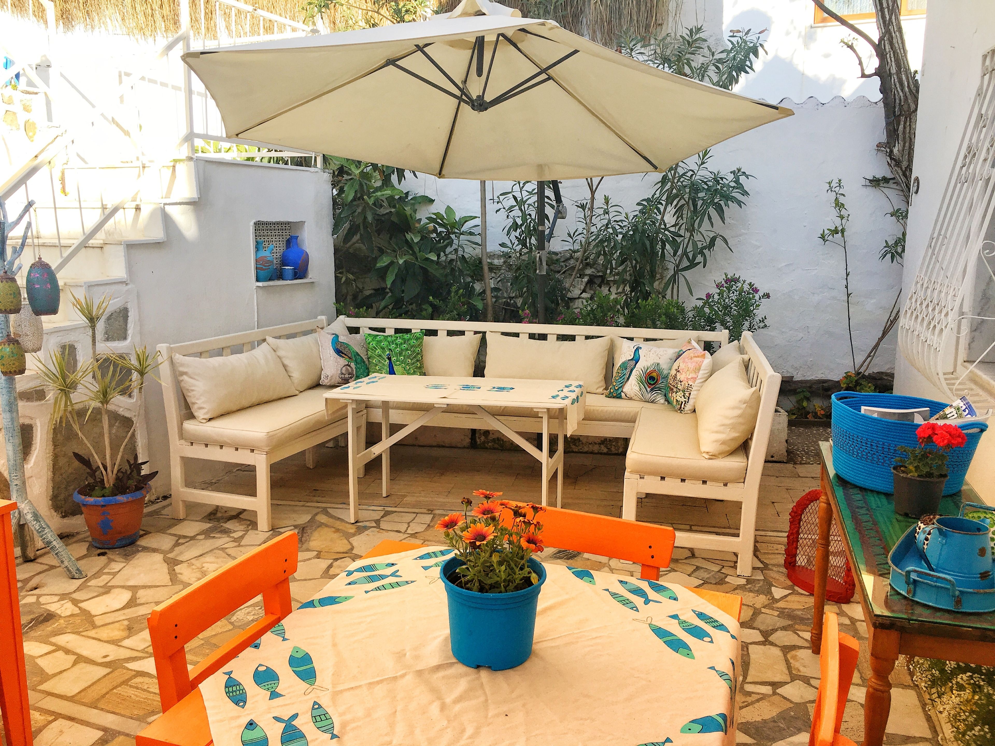 A cozy outdoor seating area with a white sofa, a table, and an umbrella. There are potted plants and a blue basket with flowers.