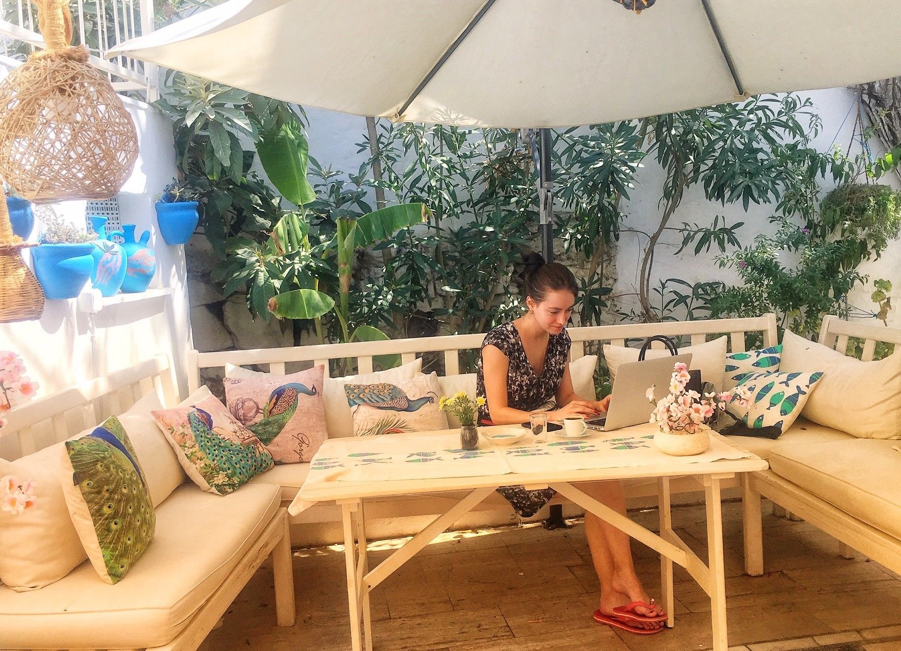 A person seated at a wooden table with a laptop, under a white umbrella, surrounded by various plants and decorative pillows.