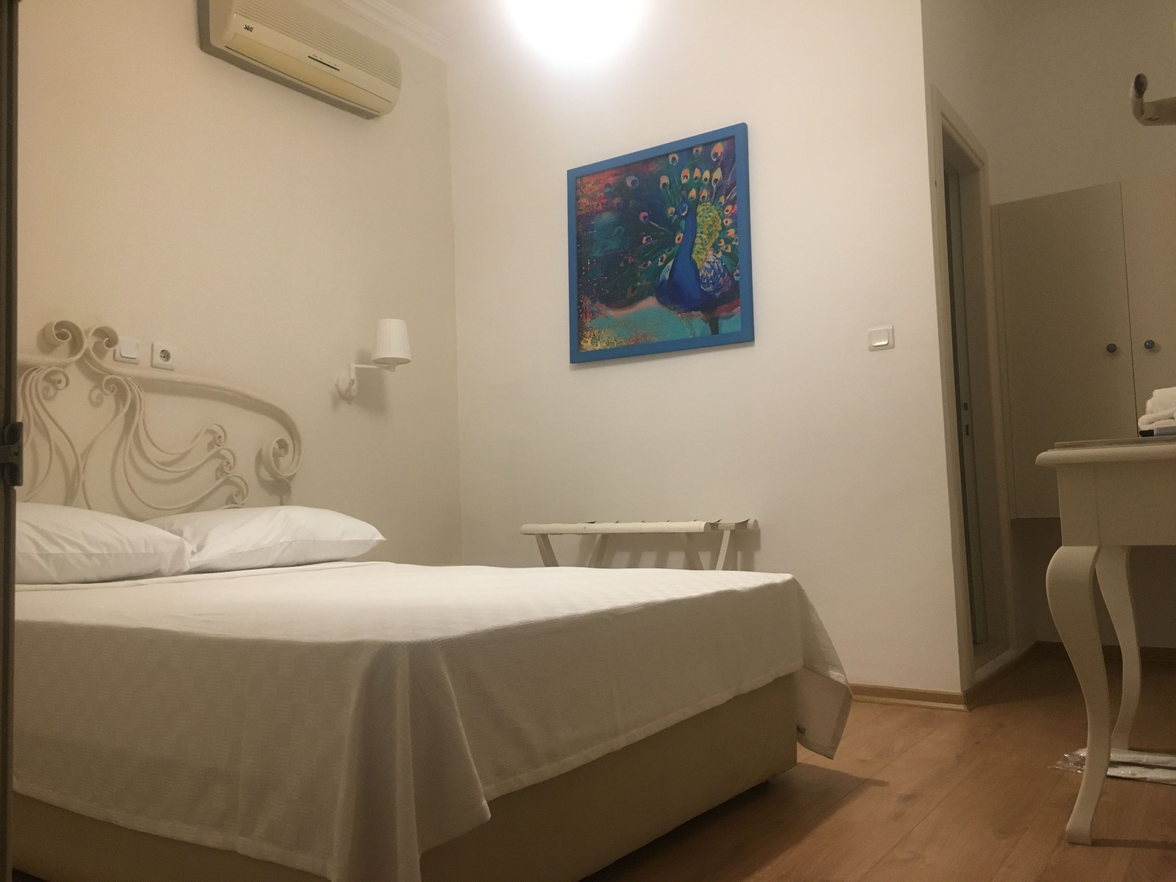 A bedroom with a bed, a nightstand, a wall-mounted air conditioner, and a painting of a peacock.