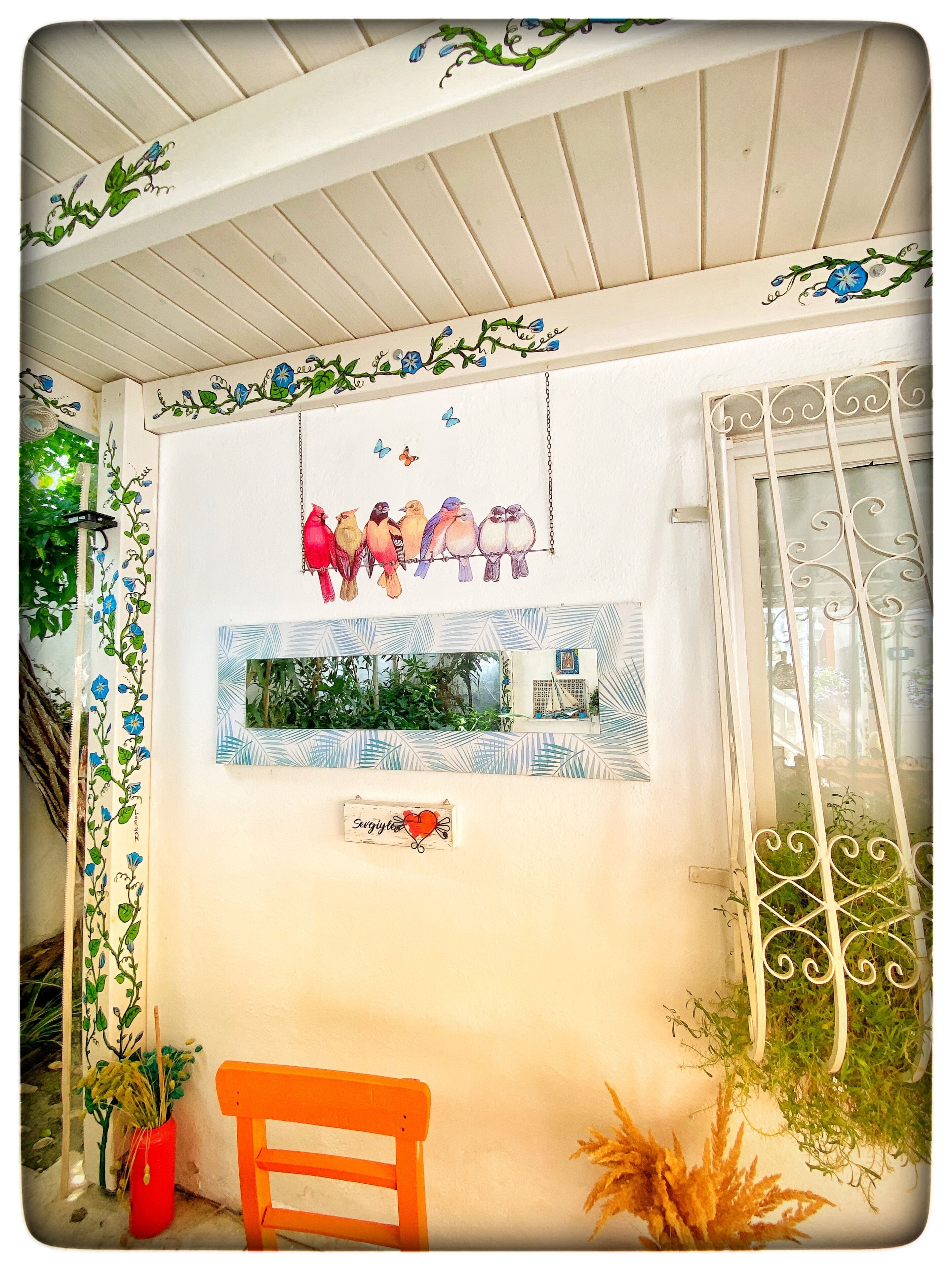 A room with a white ceiling, a window with a view of greenery, a red bench, and a wall with a bird mural and a heart-shaped sign.