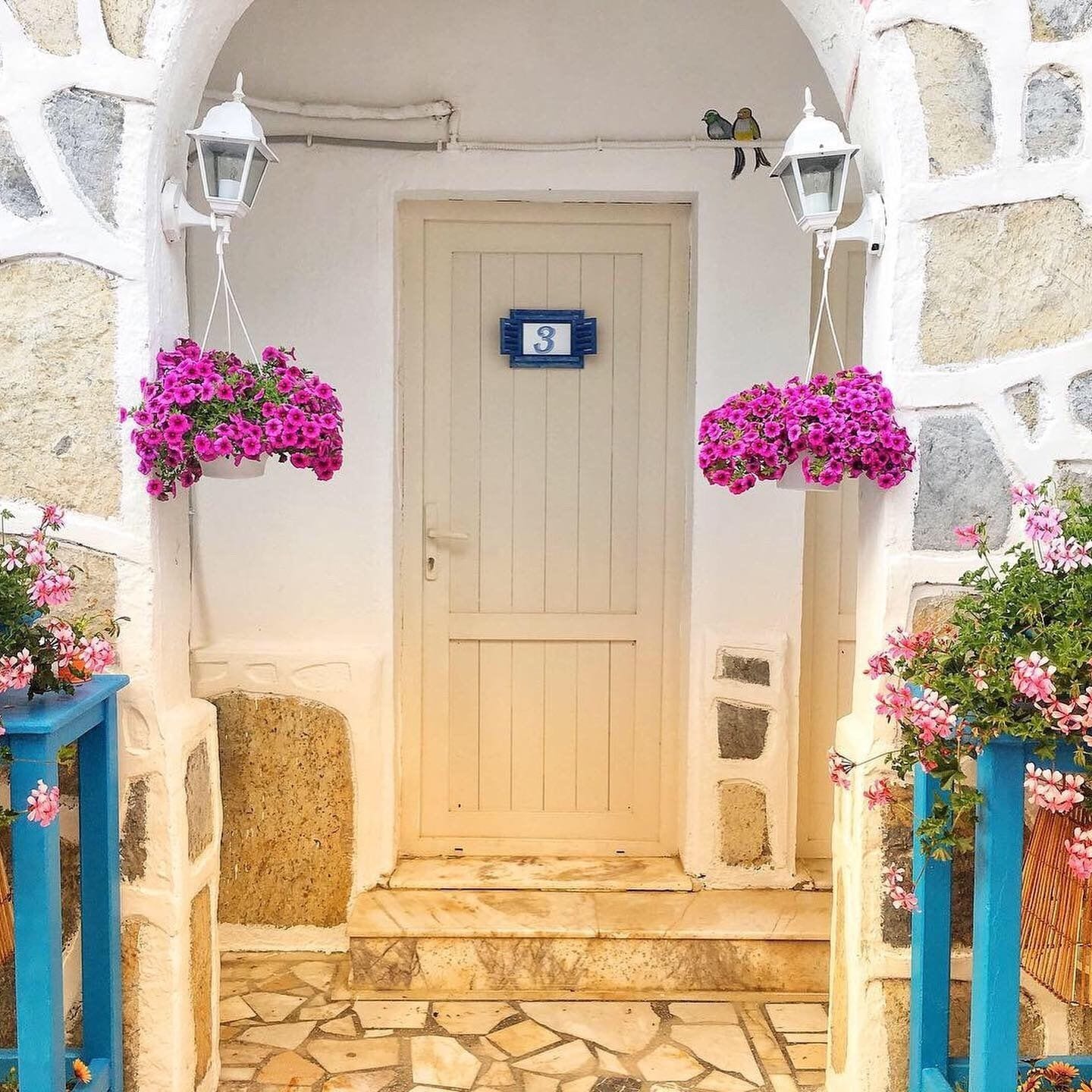 Una puerta blanca con un letrero azul en la parte superior, flanqueada por muros de piedra y cestas con flores colgantes.