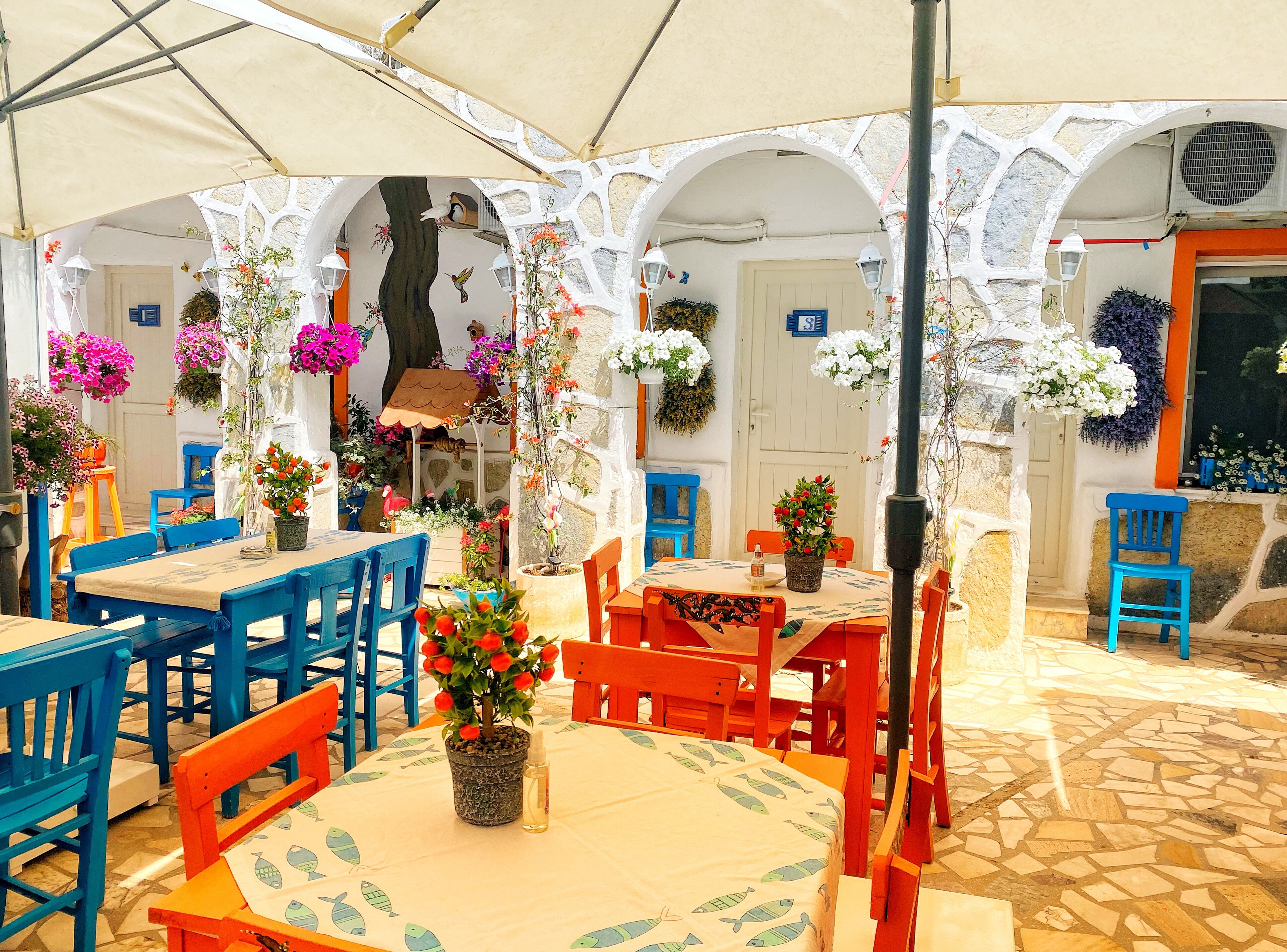 Un área de estar al aire libre con mesas y sillas, rodeada de paredes blancas y arcos, decorada con flores colgantes y plantas en macetas.