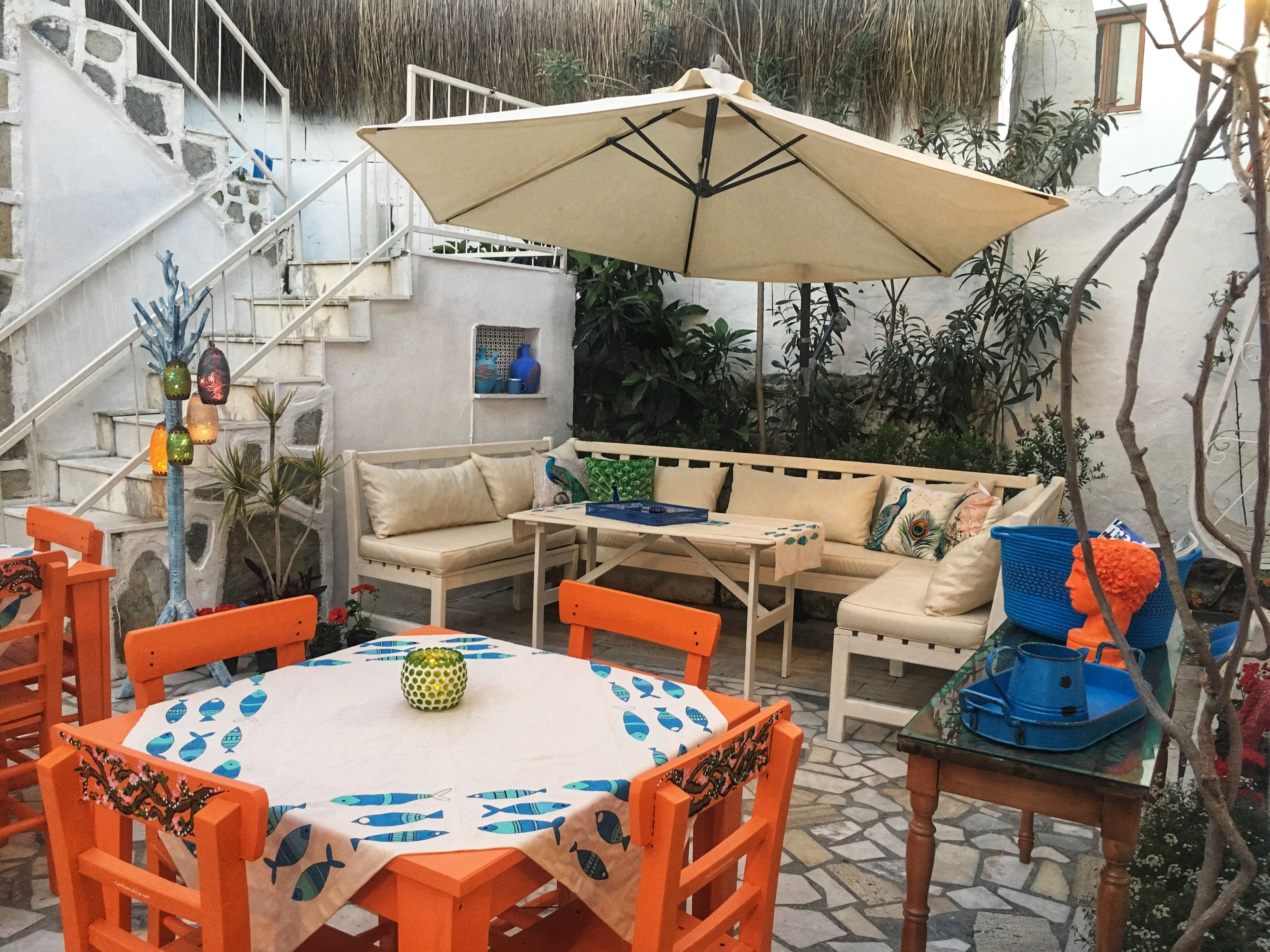Een terras met een ronde tafel, oranje stoelen, een beige bank en een grote parasol.