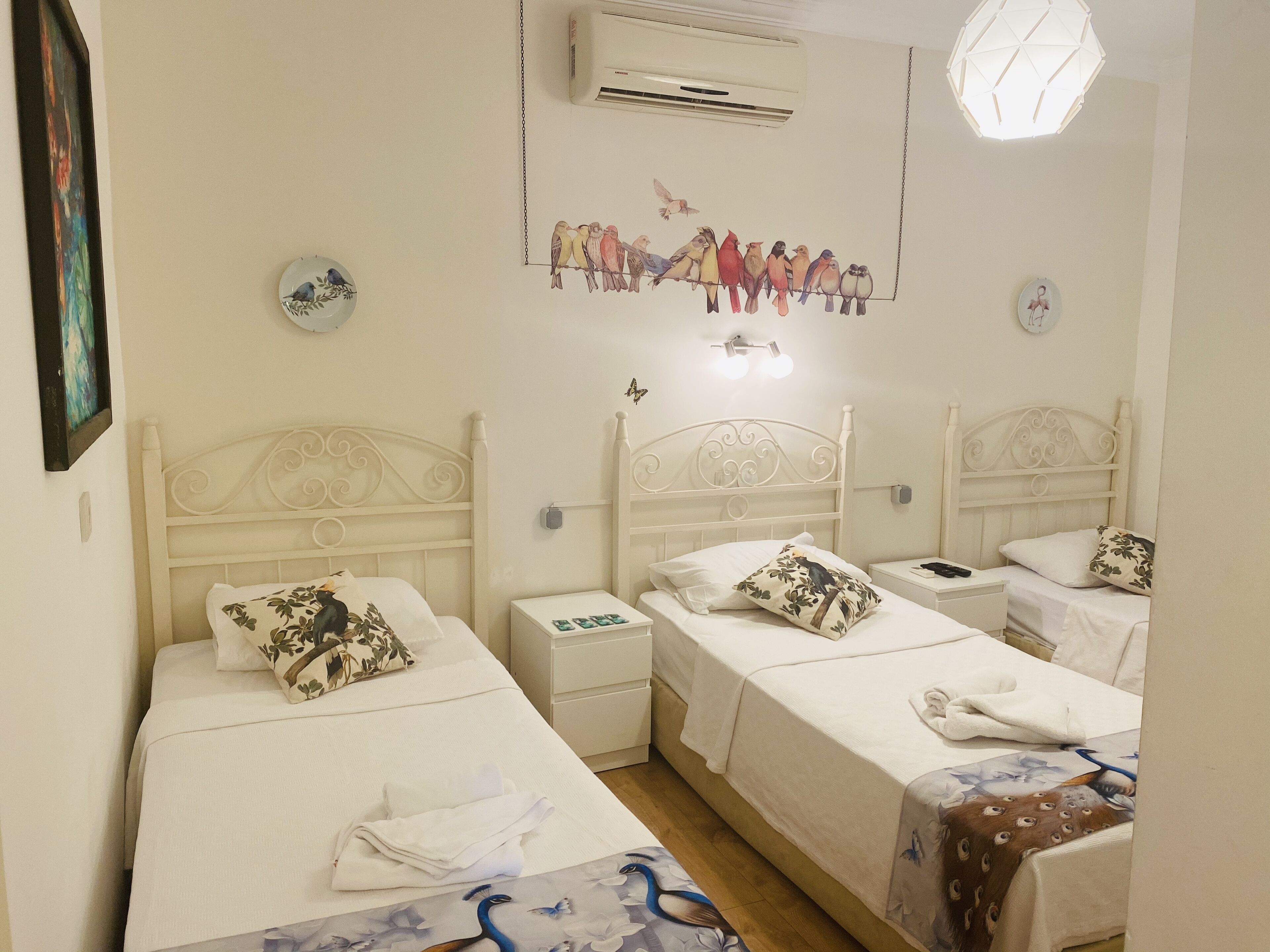 Ein Hotelzimmer mit drei Einzelbetten, jeweils mit weißem Kopfteil und passenden Nachttischen. Über den Betten ist eine wandmontierte Klimaanlage angebracht, und an der Wand ist eine Wandmalerei mit Vogelmotiv zu sehen.
