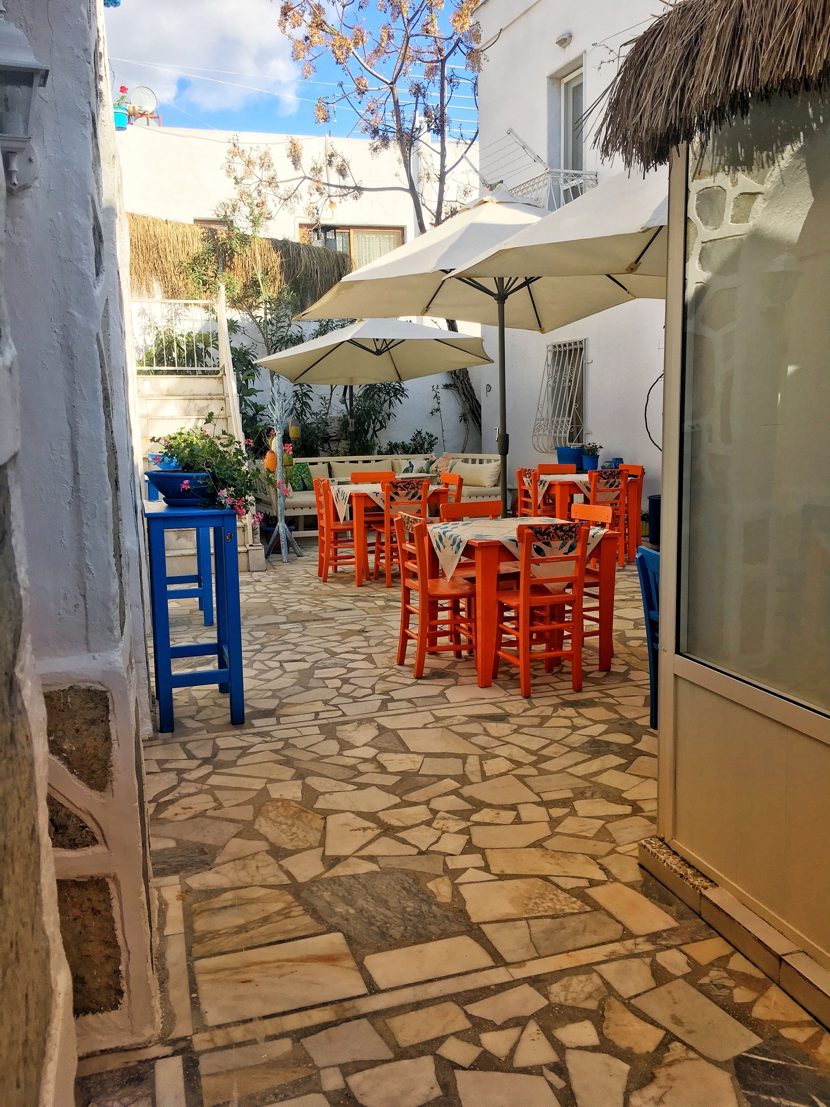 Un patio al aire libre con suelo de piedra, sillas anaranjadas y azules, y una mesa bajo un paraguas grande.
