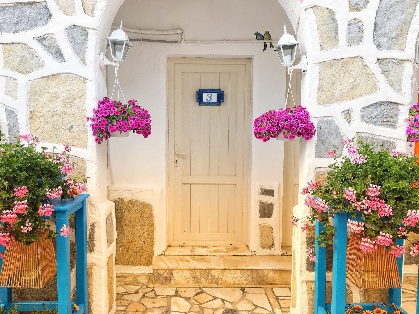 A stone-walled entrance with a wooden door, a number sign, and hanging flower baskets.