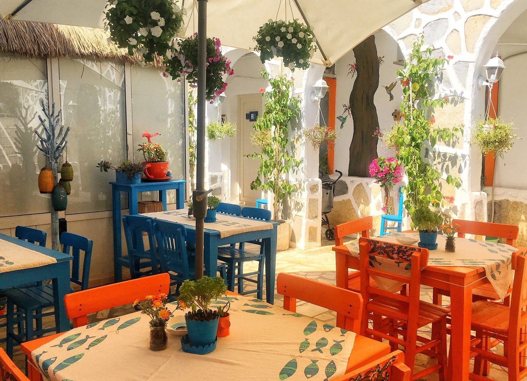 An outdoor dining area with blue and orange tables and chairs, surrounded by potted plants and hanging flower baskets.