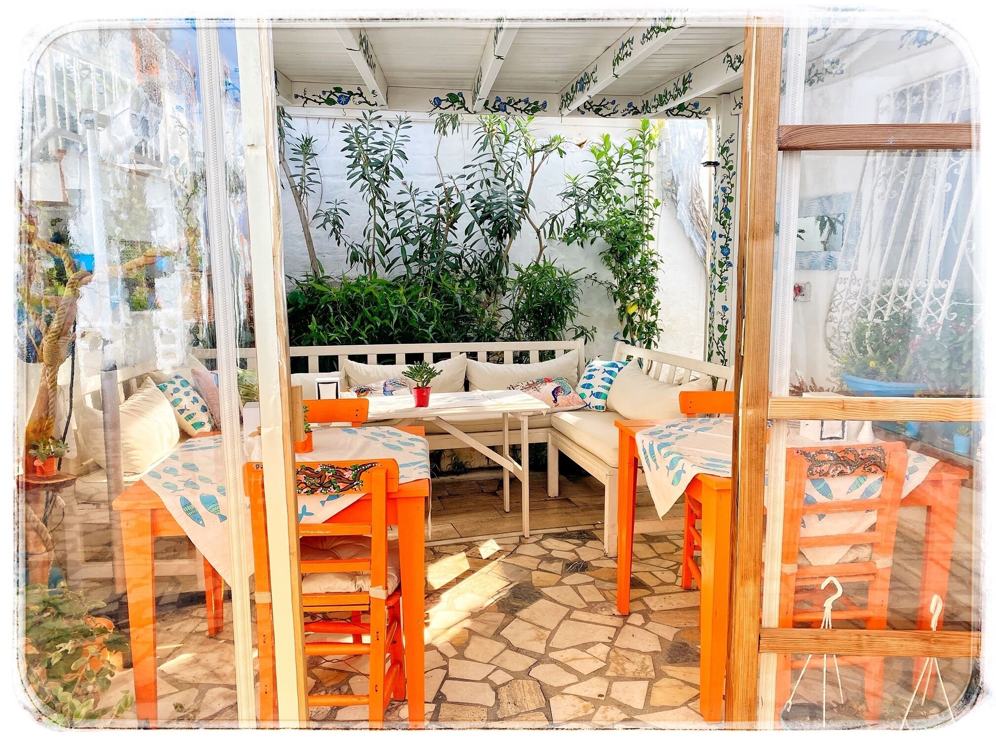 Un espace de détente extérieur accueillant, avec un banc blanc et des chaises orange, entouré d’une végétation luxuriante et doté d’un sol carrelé.