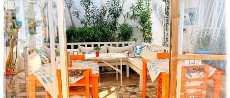 Un espace de détente extérieur accueillant, avec un banc blanc et des chaises orange, entouré d’une végétation luxuriante et doté d’un sol carrelé.