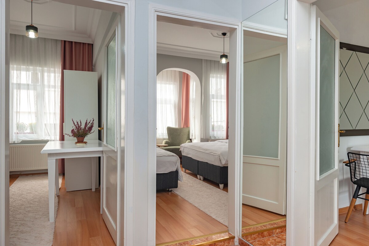 A hotel room with a white desk, two beds, a chair, and a radiator.