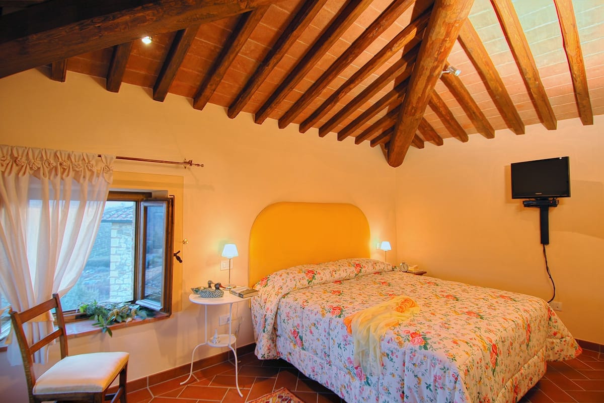 A bedroom with a wooden ceiling, a bed with floral bedding, a bedside table, a chair, and a window with curtains.