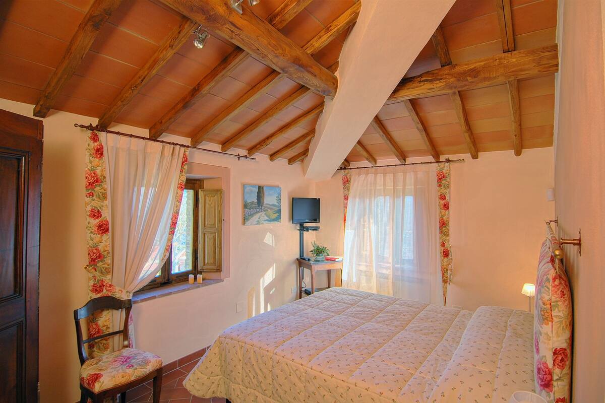 A bedroom with a wooden ceiling, a bed with floral bedding, a chair, a television, and a small table with a plant.