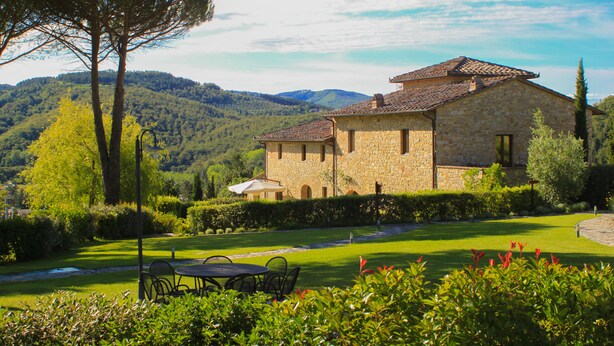 A stone building with a tiled roof, surrounded by a well-maintained garden and a view of rolling hills. - Cavarchino B&B (Gaiole in Chianti)