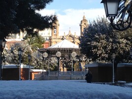 Una plaza nevada con un edificio histórico que tiene un campanario y una zona cubierta con faroles decorativos.