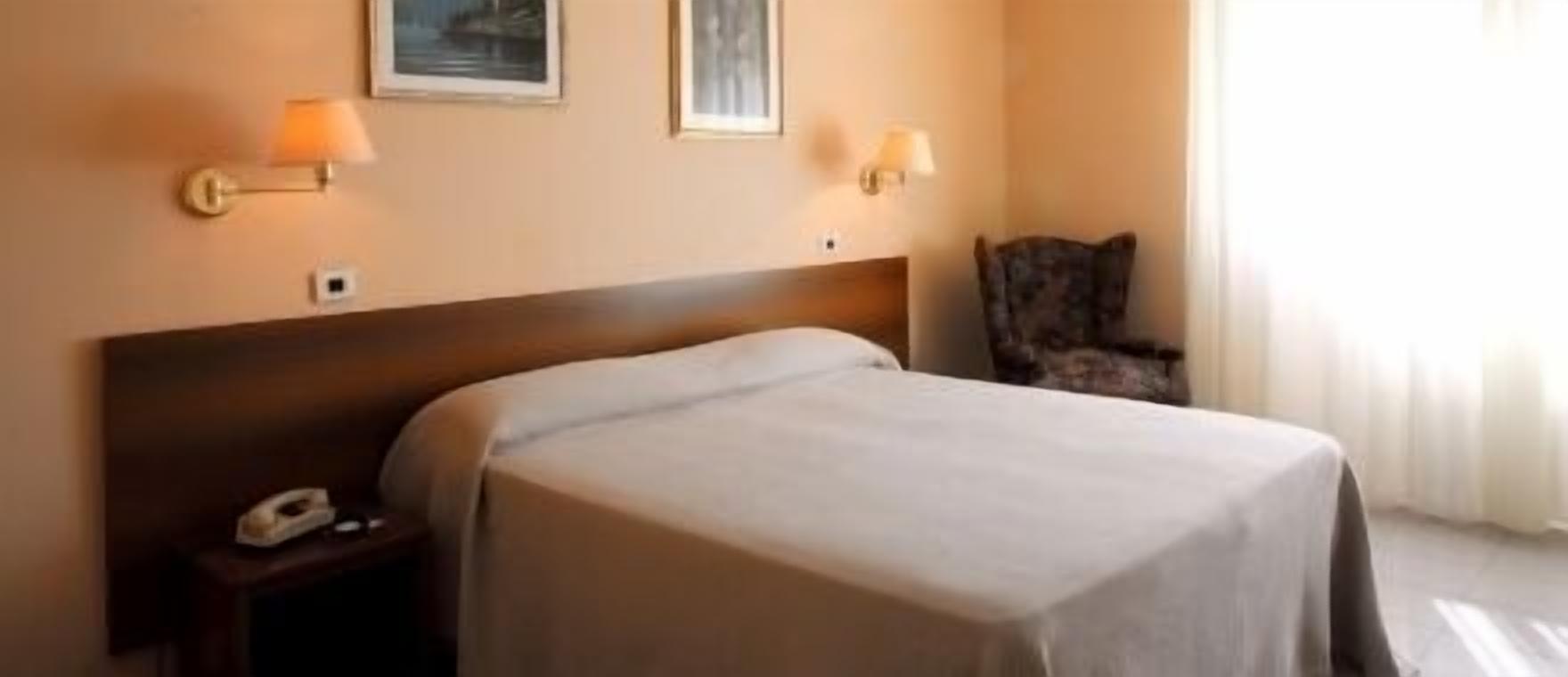 A hotel room with a bed, a nightstand with a telephone, two wall-mounted lamps, and a chair.