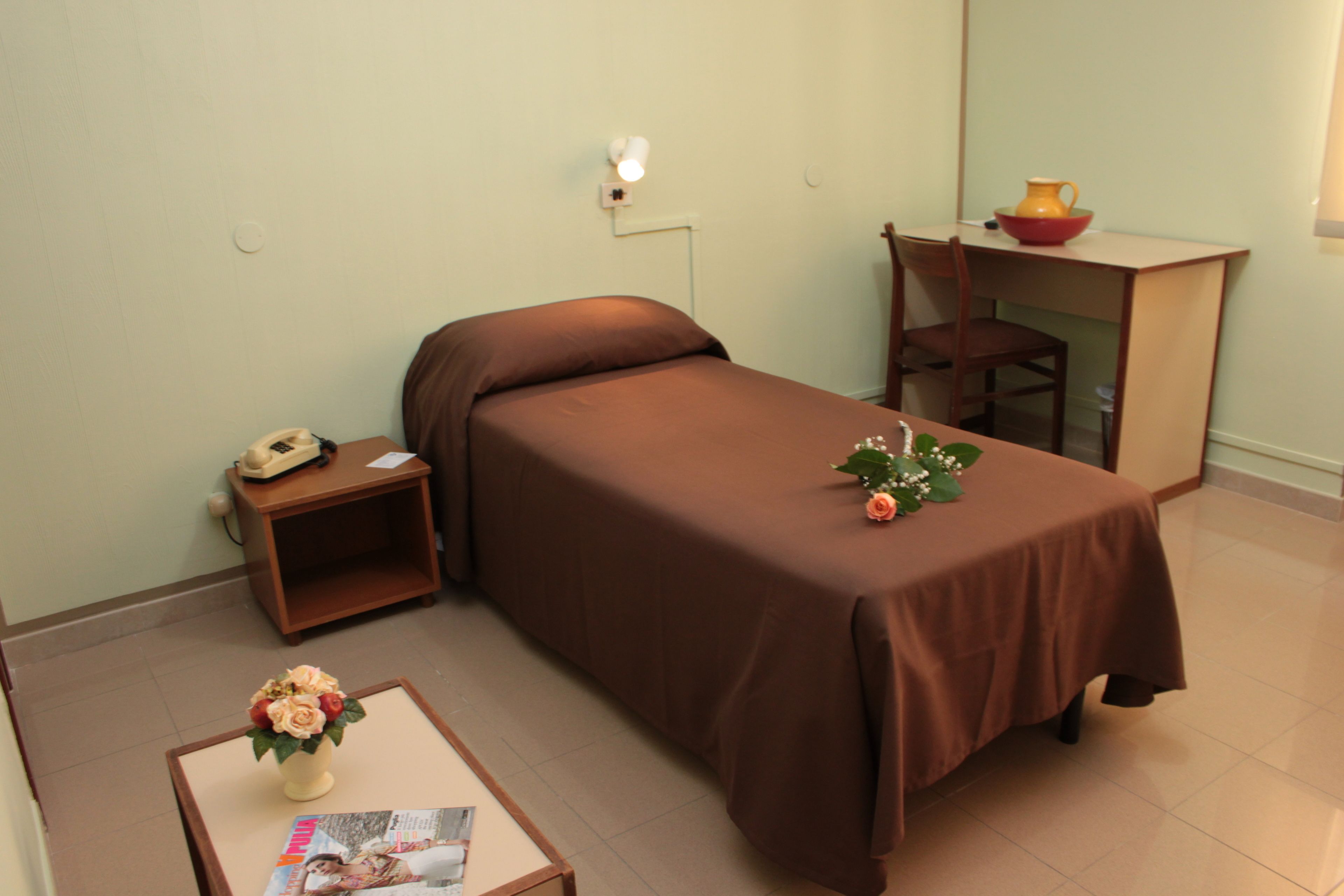 A small, single-bed room with a brown bedspread, a bedside table with a lamp, a chair, and a small table with a vase of flowers.