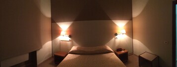 Una habitación de hotel estrecha con una cama centrada entre dos mesitas de noche, cada una con una lámpara.