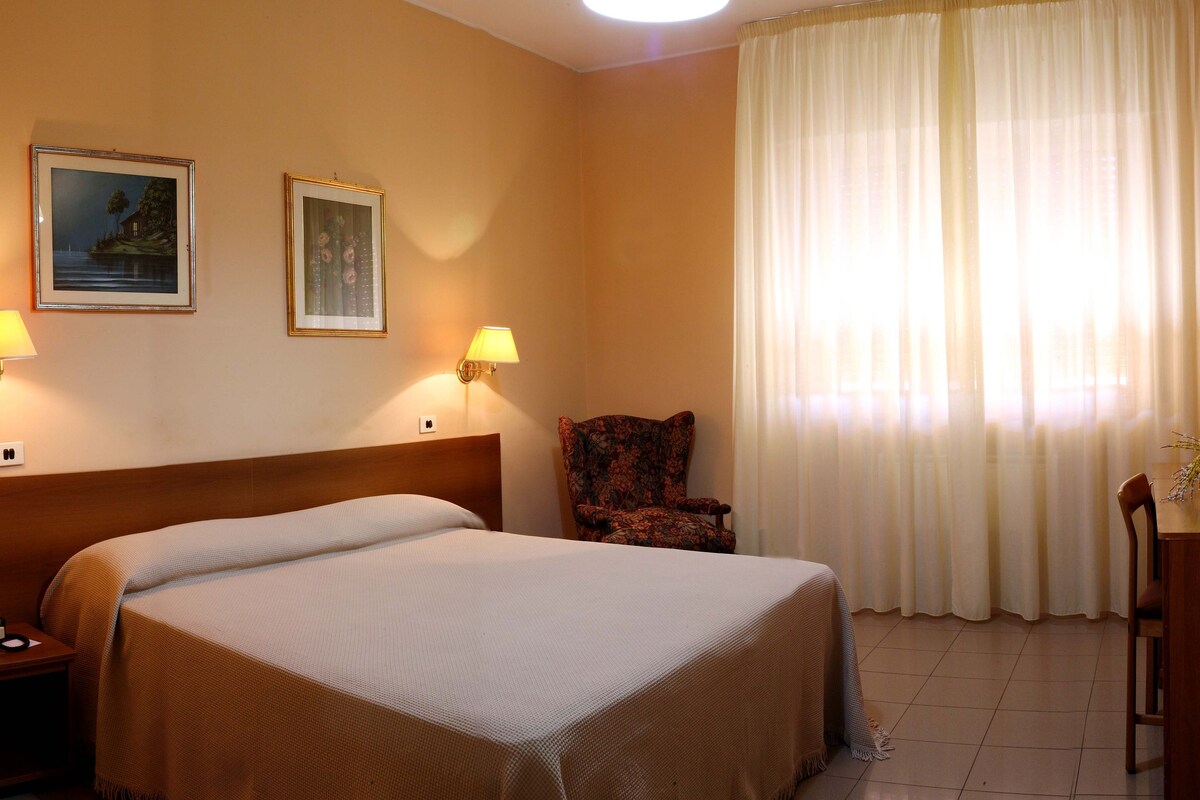 Habitación de hotel con cama, dos lámparas de pared, una silla, escritorio con frutero y televisor en la pared.