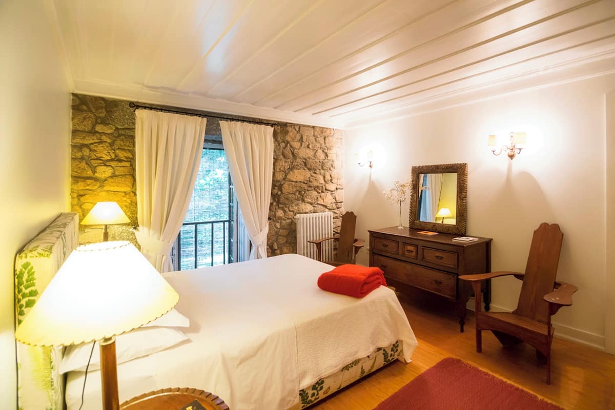 Une chambre avec un mur en pierre, un lit avec une serviette rouge, une commode en bois, une chaise et une lampe.