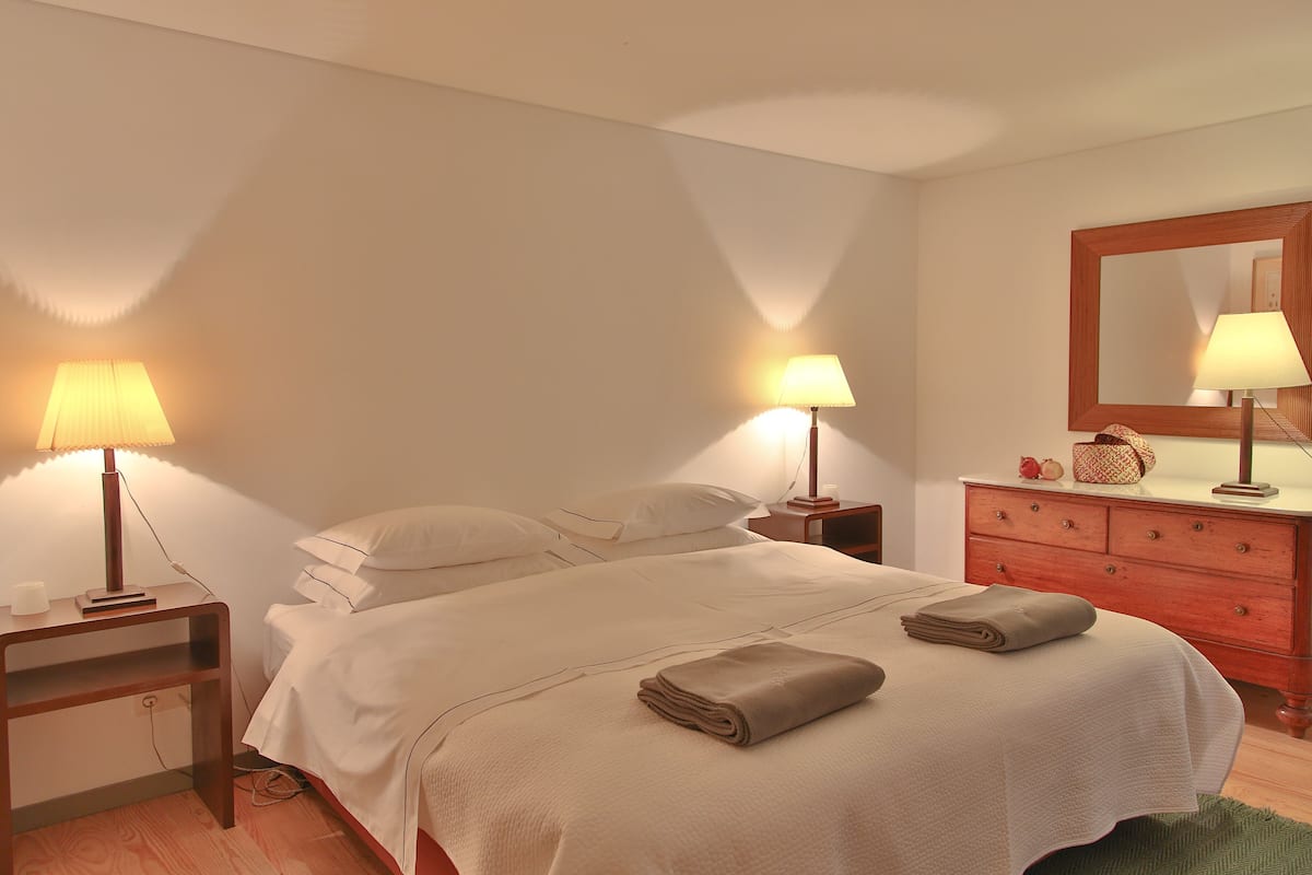 Un lit bien fait, avec une couverture pliée, deux lampes de chevet, une table de nuit en bois, une commode avec un miroir et un tapis vert.