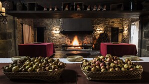 Een knus vertrek met een stenen open haard, twee rode tafels en manden vol fruit.