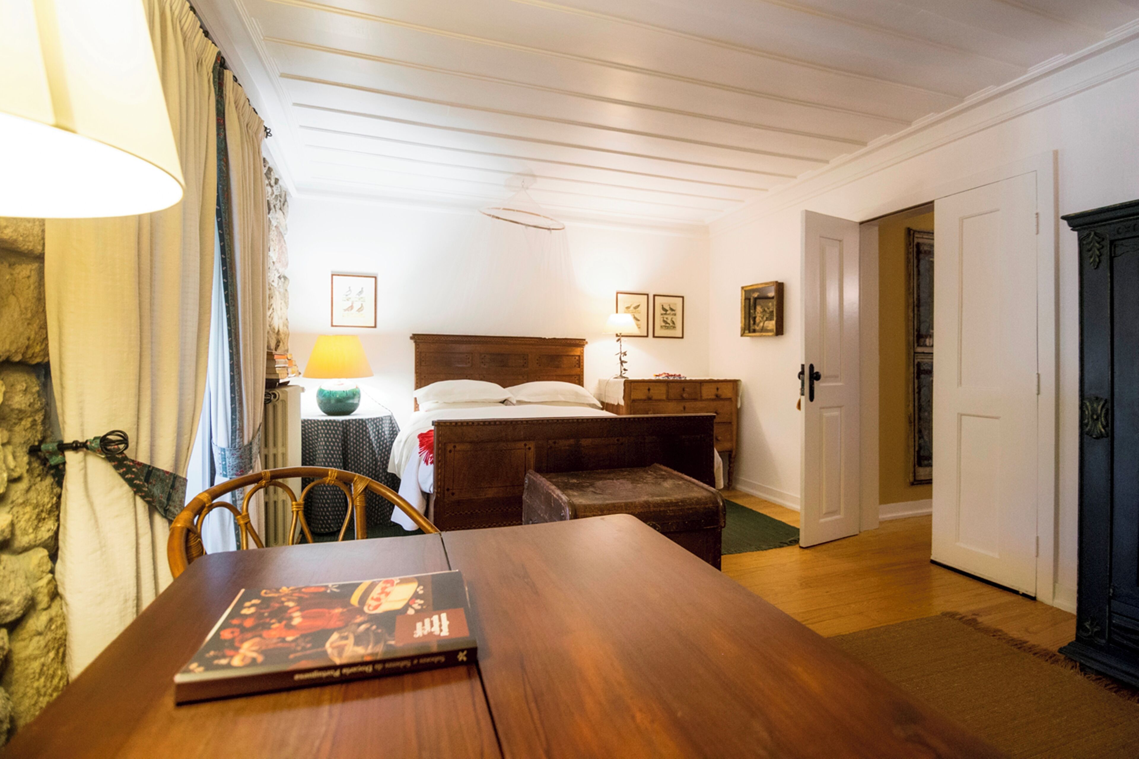 Een kamer met een houten tafel, een bed met een houten hoofdeinde, een nachtkastje met een lamp en een stenen muur.
