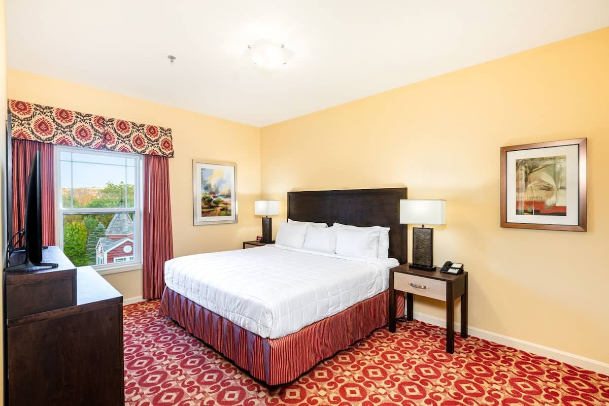 A hotel room with a bed, two bedside tables, a desk, a window with curtains, and two framed pictures on the walls.