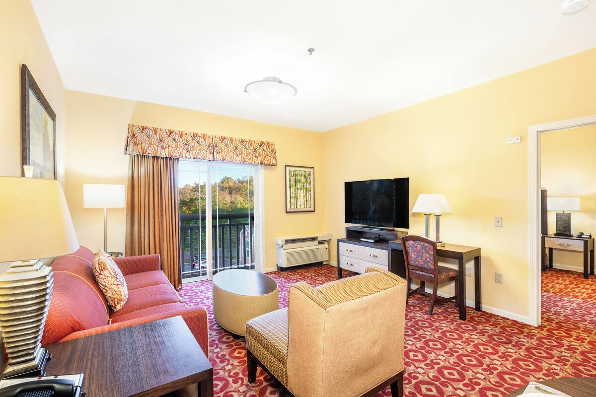 Habitación de hotel con sofá, sillón, televisión, escritorio y un amplio ventanal con cortinas.